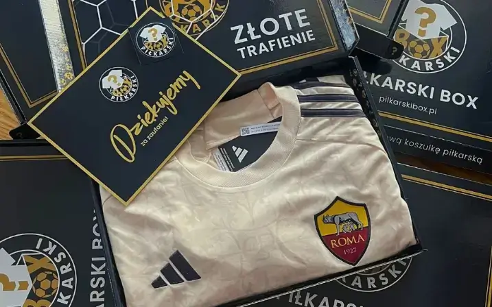 Otwarty Mystery Box z koszulką AS Roma w kolorze beżowym i gratisami w Boxie. W tle kartoniki PIŁKARSKI BOX.